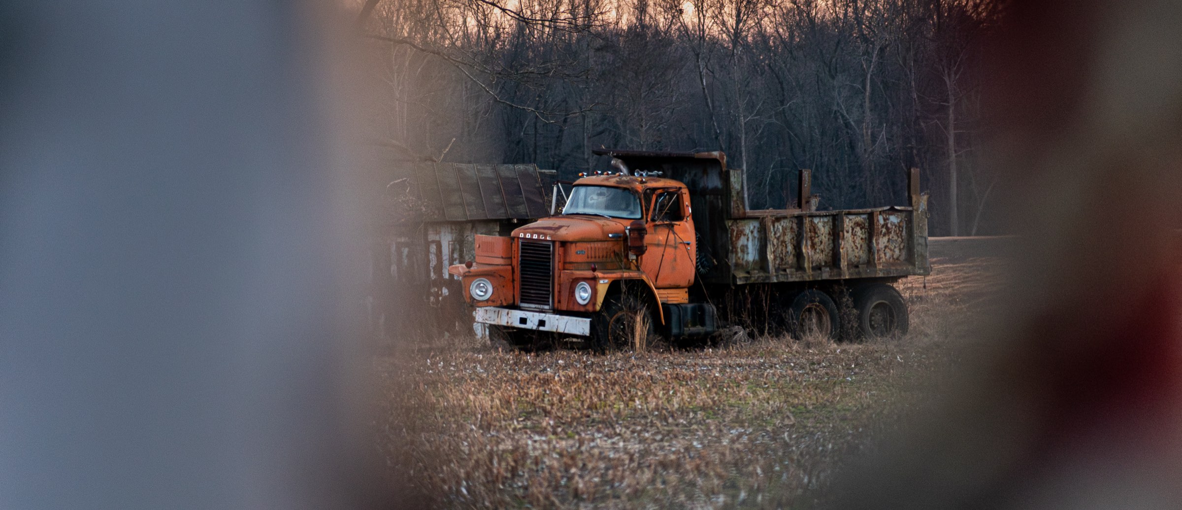 An old rusted Dodge dump truck sitting in a field at sunset, framed by bare winter trees.